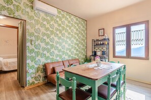 Una habitación con una mesa de madera, sillas verdes, un sofá de cuero marrón, un papel tapiz estampado y una ventana con cortinas.