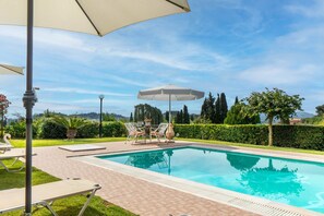 Piscina com espreguiçadeiras e um guarda-sol, cercada por vegetação e céu claro.