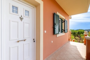Una puerta blanca con picaporte y ventana con contraventanas, un balcón con suelo de baldosas, y vista a zonas verdes.