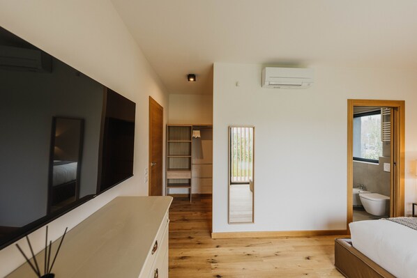A bedroom with a large flat-screen TV, wooden flooring, a bed, a wardrobe, and a bathroom visible through an open door.