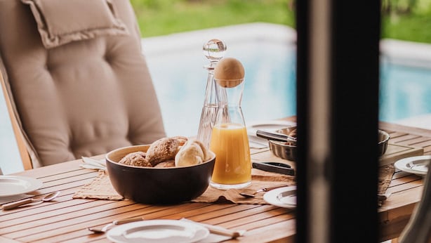 Ein Holztisch mit einer Schüssel Kekse, einer Glaskaraffe und Tellern, vor dem Hintergrund eines Schwimmbeckens und Grünflächen.