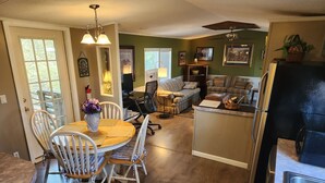 A dining area with a wooden table and white chairs, a kitchen with a refrigerator, and a living room with a sofa and chairs.