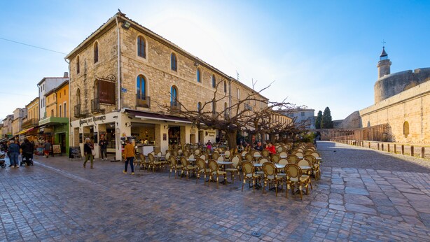 A cobblestone street with outdoor seating, a stone building with a balcony, and a tower in the background. - 'Petite Maison Aigues Mortes' with Sea View, Shared Pool and Wi-Fi (Aigues-Mortes)