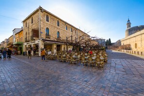 A cobblestone street with outdoor seating, a stone building with a balcony, and a tower in the background. - 'Petite Maison Aigues Mortes' with Sea View, Shared Pool and Wi-Fi (Aigues-Mortes)