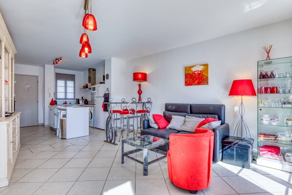 A modern living room with a sofa, a glass coffee table, and a red chair. There is a kitchen area in the background with a dining table and chairs.