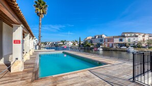 A swimming pool with a wooden deck, a palm tree, and buildings in the background. - 'Petite Maison Aigues Mortes' with Sea View, Shared Pool and Wi-Fi (Aigues-Mortes)