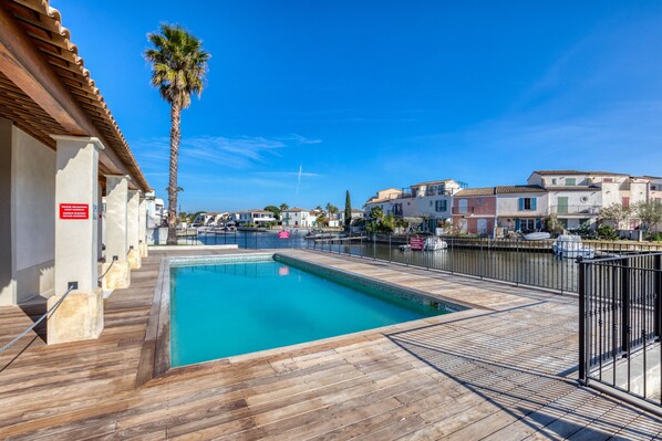 A swimming pool with a wooden deck, a palm tree, and buildings in the background.