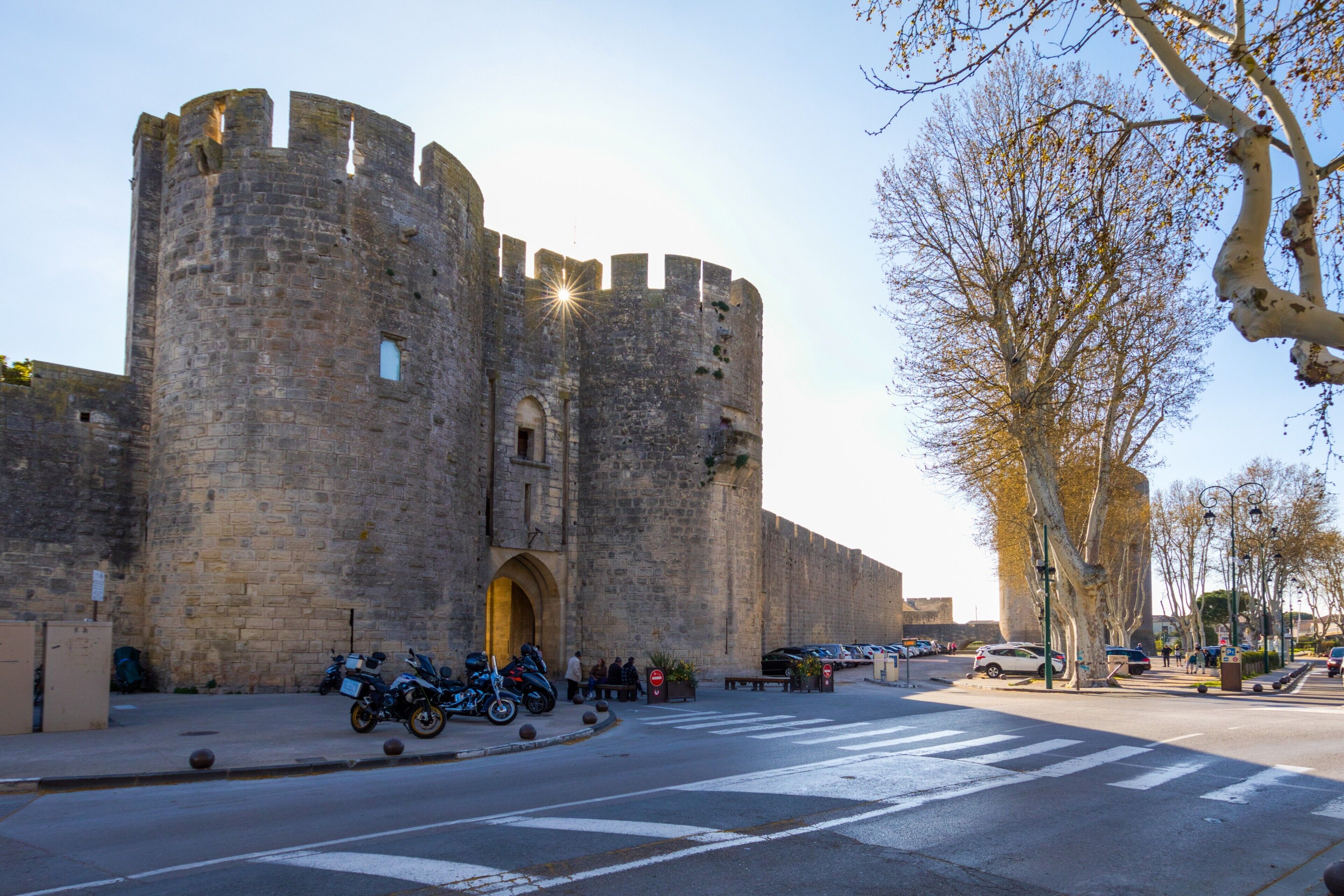 A historic stone castle with a large arched entrance, parked motorcycles, and a clear sky.