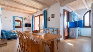 Una mesa de comedor de madera con sillas, una cocina con armarios azules y una sala de estar con un sofá y una planta.