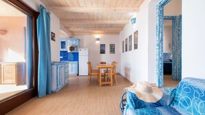 Una habitación con techo de madera, una mesa de comedor con sillas, un sofá, una zona de cocina con armarios y electrodomésticos, y una ventana con cortinas.