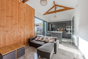 Une cuisine moderne avec des meubles de couleur sombre et des appareils en acier inoxydable, un banc en bois et un canapé d’angle gris avec des coussins.