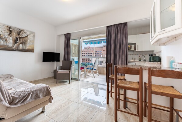 A modern hotel room with a bed, a TV, a dining area with chairs, a kitchen area, and a balcony with a view of buildings.