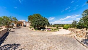 A paved driveway leading to a building with a stone wall on one side and a tree-lined area with a statue. - 'Villa Sunrise' with Shared Terrace, Wi-Fi, and AC – Ideal for 4/5 Adults and 1 Child (Gallipoli)