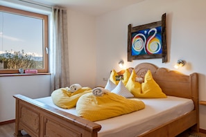A bed with yellow bedding, a wooden headboard, a painting on the wall, and a window with a view of mountains.