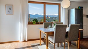Un comedor con mesa, sillas, una ventana con vistas a montañas, un refrigerador y una planta en maceta.