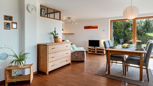 Un salón moderno con suelo de madera, una mesa de comedor con sillas, un sofá, un televisor y un amplio ventanal con vistas a zonas verdes.