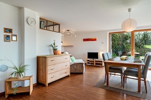Un salon moderne avec un plancher en bois, une table à manger avec des chaises, un canapé, un téléviseur et une grande fenêtre donnant sur un paysage verdoyant.