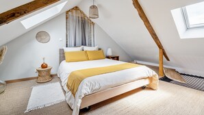 A bedroom with a sloped ceiling, a bed with a white and yellow bedspread, a woven rug, a small side table, and a window. - "Le Kerhuel d'Agnès" with private terrace, private garden and Wi-Fi (Plouasne)