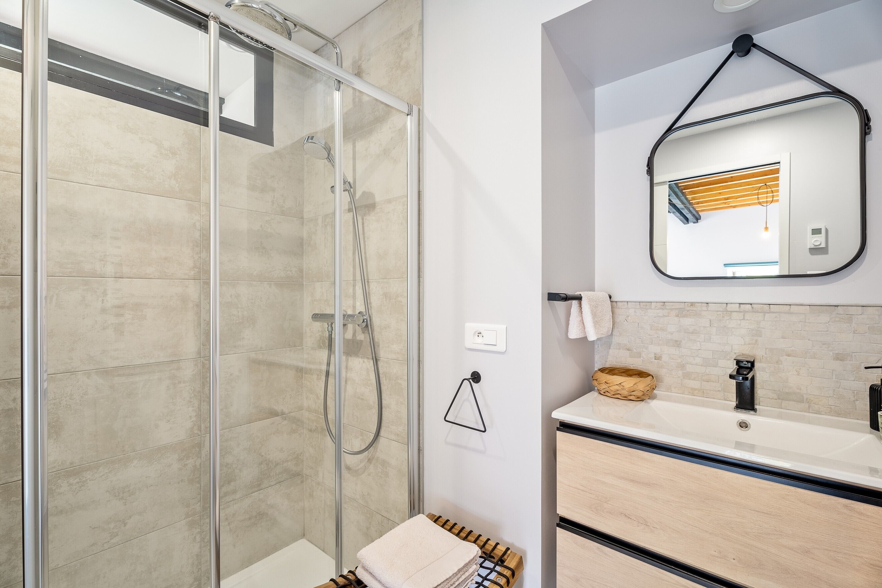 Un bany modern amb plat de dutxa de vidre, un lavabo amb paraire i un mirall amb estoreta de fusta.