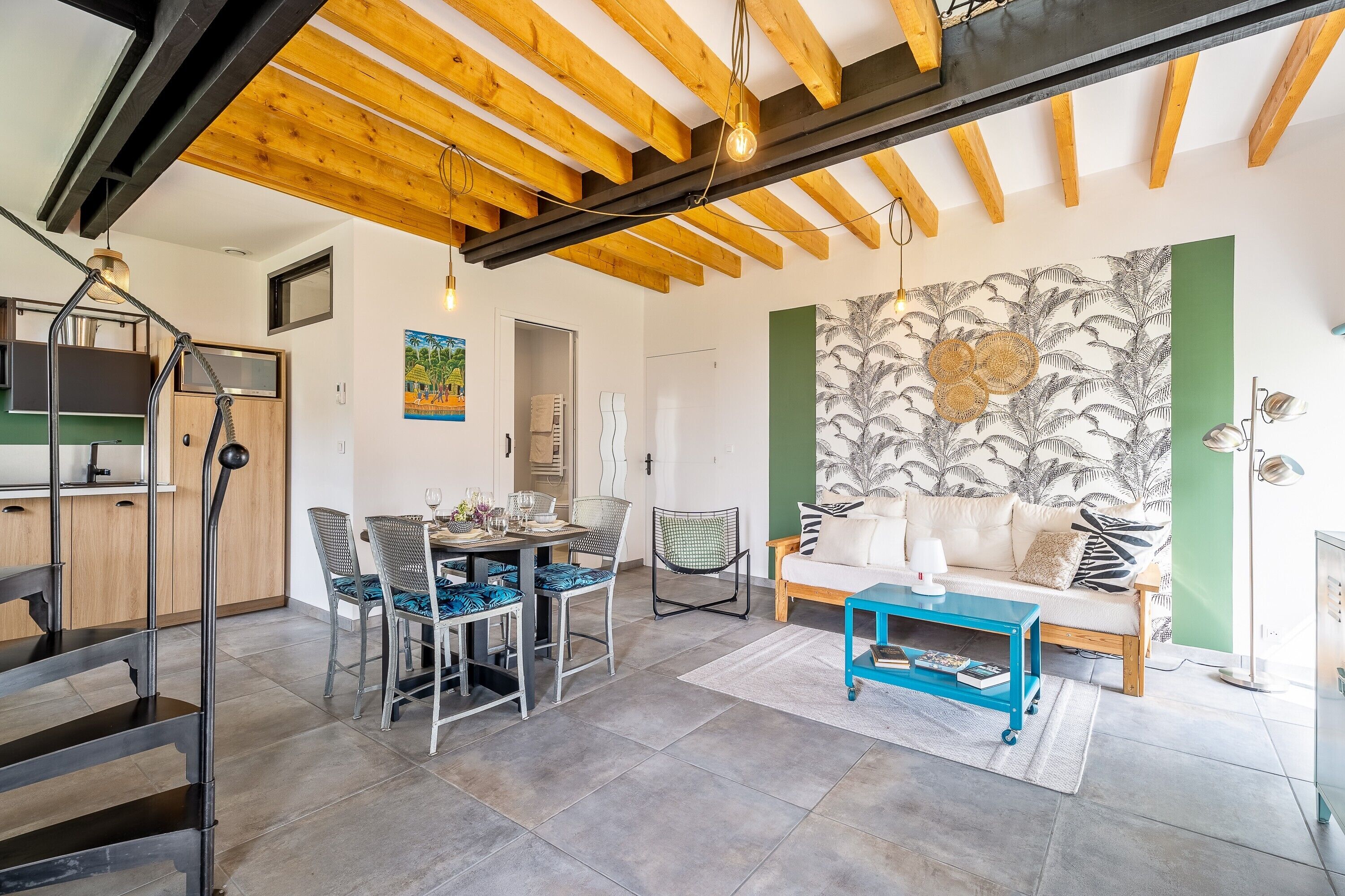 Un intérieur moderne avec des poutres apparentes, un coin repas avec des chaises et une table, un canapé avec des coussins et un mur orné d’un motif de feuilles tropicales.
