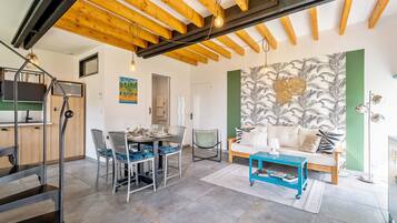 Un intérieur moderne avec des poutres apparentes, un coin repas avec des chaises et une table, un canapé avec des coussins et un mur orné d’un motif de feuilles tropicales.