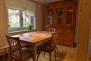 A dining area with a wooden table and chairs, a window with blinds, and a wooden cabinet with glass doors. - Holiday Home "Chez Jeanne, Charming House" with Private Garden and Wi-Fi (Dossenheim-sur-Zinsel)
