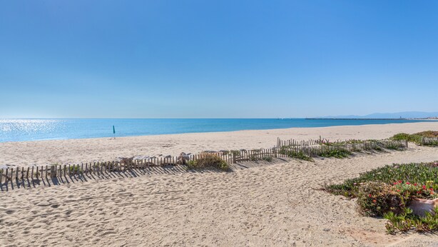 A sandy beach with a wooden fence and some greenery. - Naturist holiday home 'Résidence Naturiste Eden' with sea view, private terrace, and Wi-Fi (Leucate)