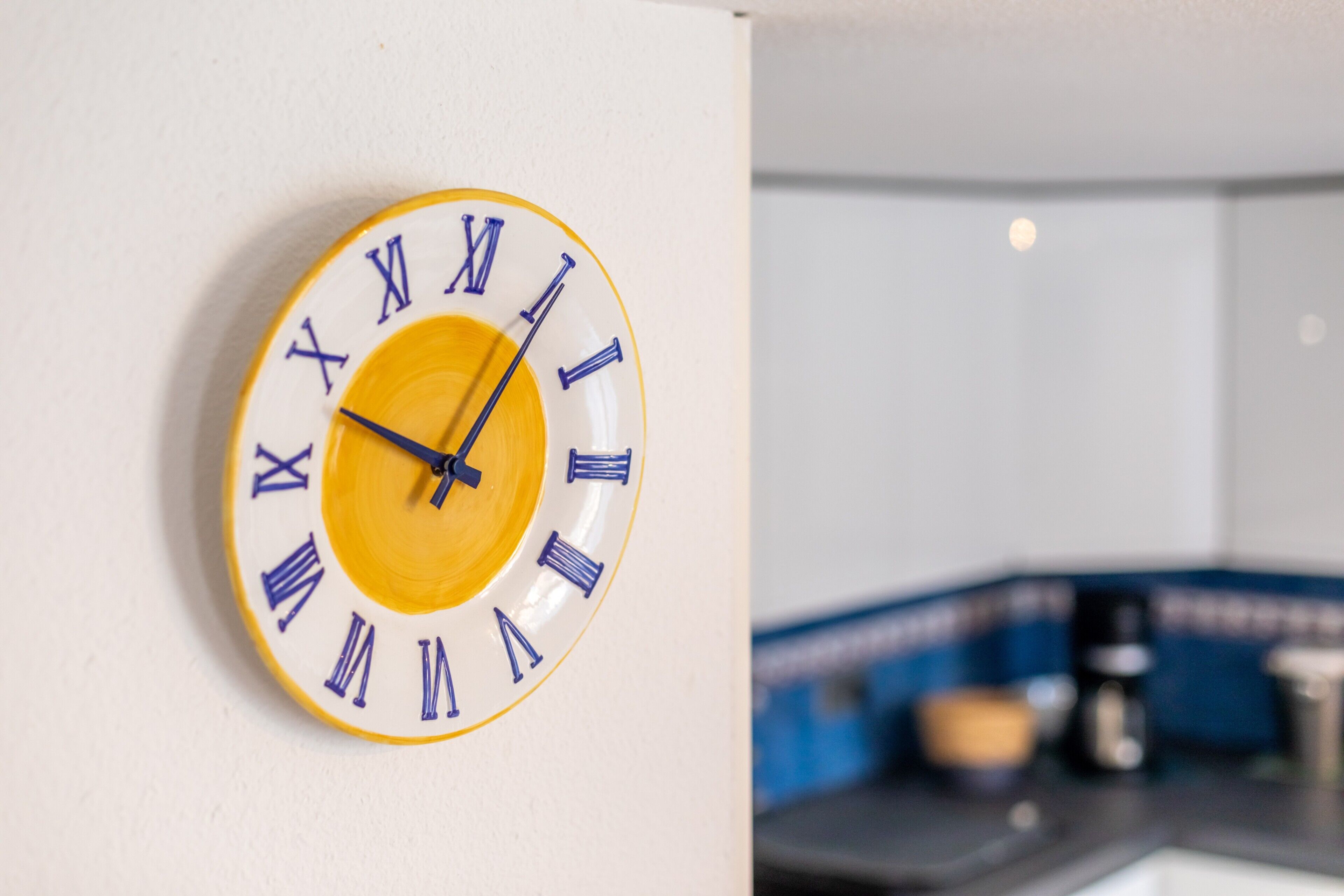 A wall-mounted clock with Roman numerals, a kitchen area with a coffee machine, and a blue countertop.