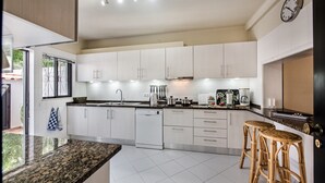 Een moderne keuken met witte kasten, een granieten aanrecht, een vaatwasser en twee rieten barstoelen.