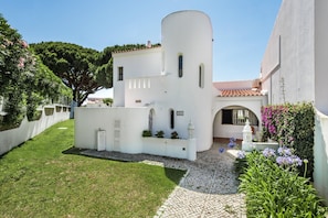 A white building with a curved tower, a paved courtyard, and a small garden with flowers.