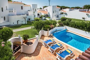 A poolside area with lounge chairs, a barbecue grill, and a view of white buildings with red-tiled roofs.