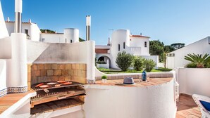 Une cuisine extérieure équipée d’un barbecue, avec des murs blancs et une vue sur un bâtiment blanc doté d’un toit rouge.