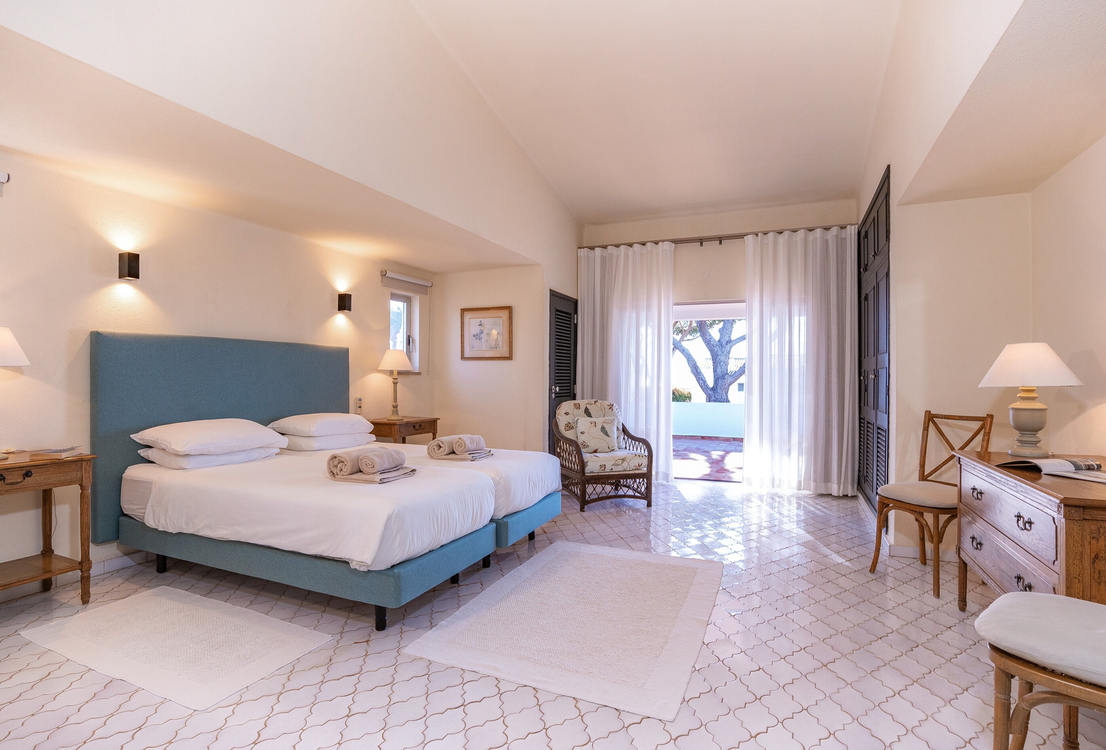 Une chambre à coucher comprenant un lit, deux tables de chevet, une chaise, un bureau et une fenêtre avec des rideaux.