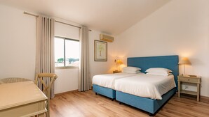 Ein Schlafzimmer mit Bett, Sessel, Tisch, einem Fenster mit Vorhängen, einer Wand-Klimaanlage und einem gerahmten Bild.