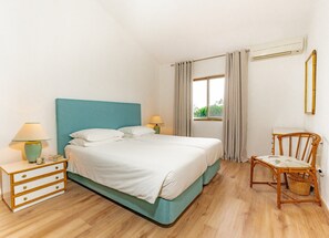 Une chambre à coucher comprenant un lit, deux tables de chevet, une chaise, une fenêtre avec des rideaux et un appareil de climatisation.