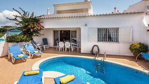 Un espace piscine avec des chaises longues et une table, un bâtiment blanc avec un toit en tuiles et un ciel dégagé.