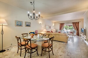 A dining area with a round table, chairs, and a chandelier. There is a sofa and a television in the background. - Villa "Mng Elliott 49" with Private Pool & Wi-Fi (Vale do Lobo)
