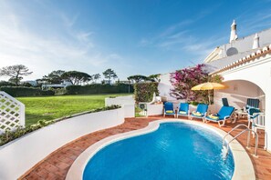 Una piscina con agua azul, rodeada de paredes blancas y un patio de ladrillo. Hay sillas reclinables azules y un paraguas amarillo.