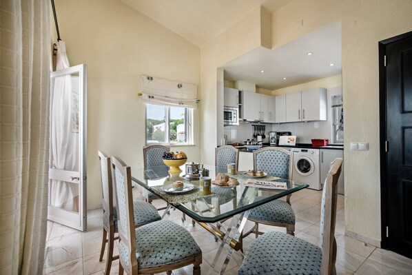 A dining area with a glass table, patterned chairs, and a view into the kitchen.