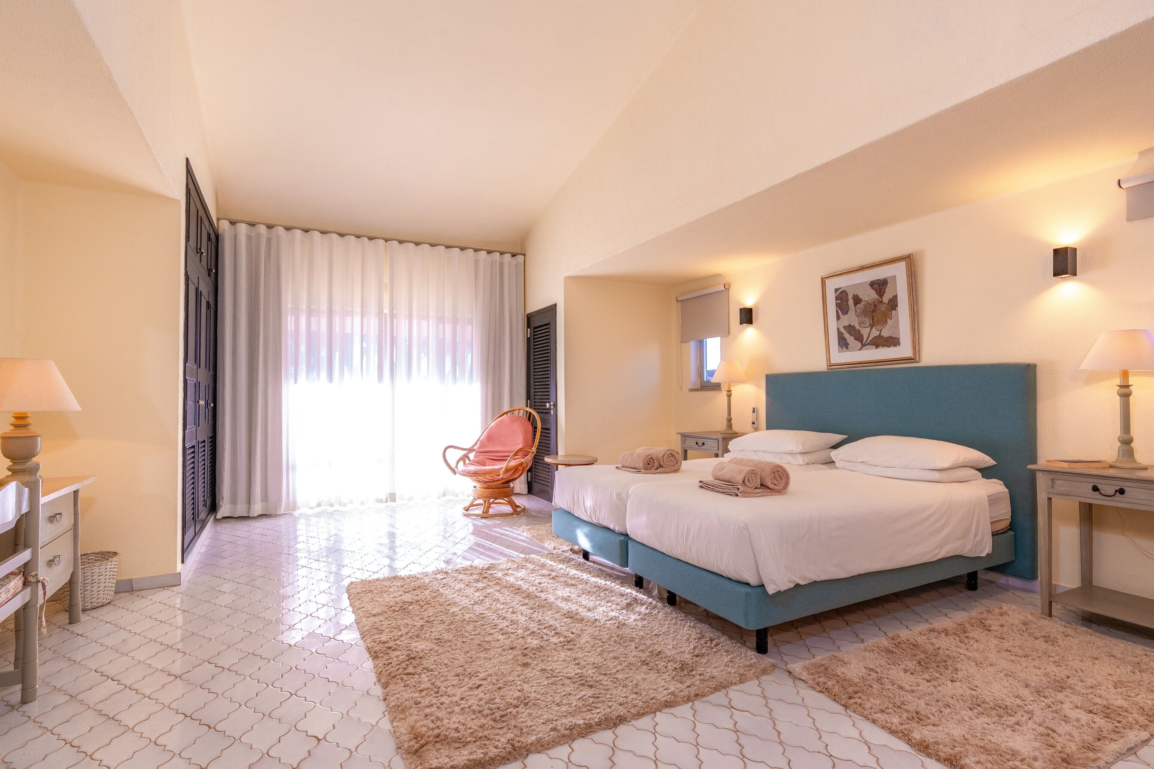 Een slaapkamer met een bed, een stoel, een nachtkastje, een lamp en een raam met gordijnen.