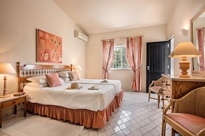 Un dormitorio con dos camas, cabeceras de madera, una mesita de noche con lámpara, una silla, un aparador y una ventana con cortinas.