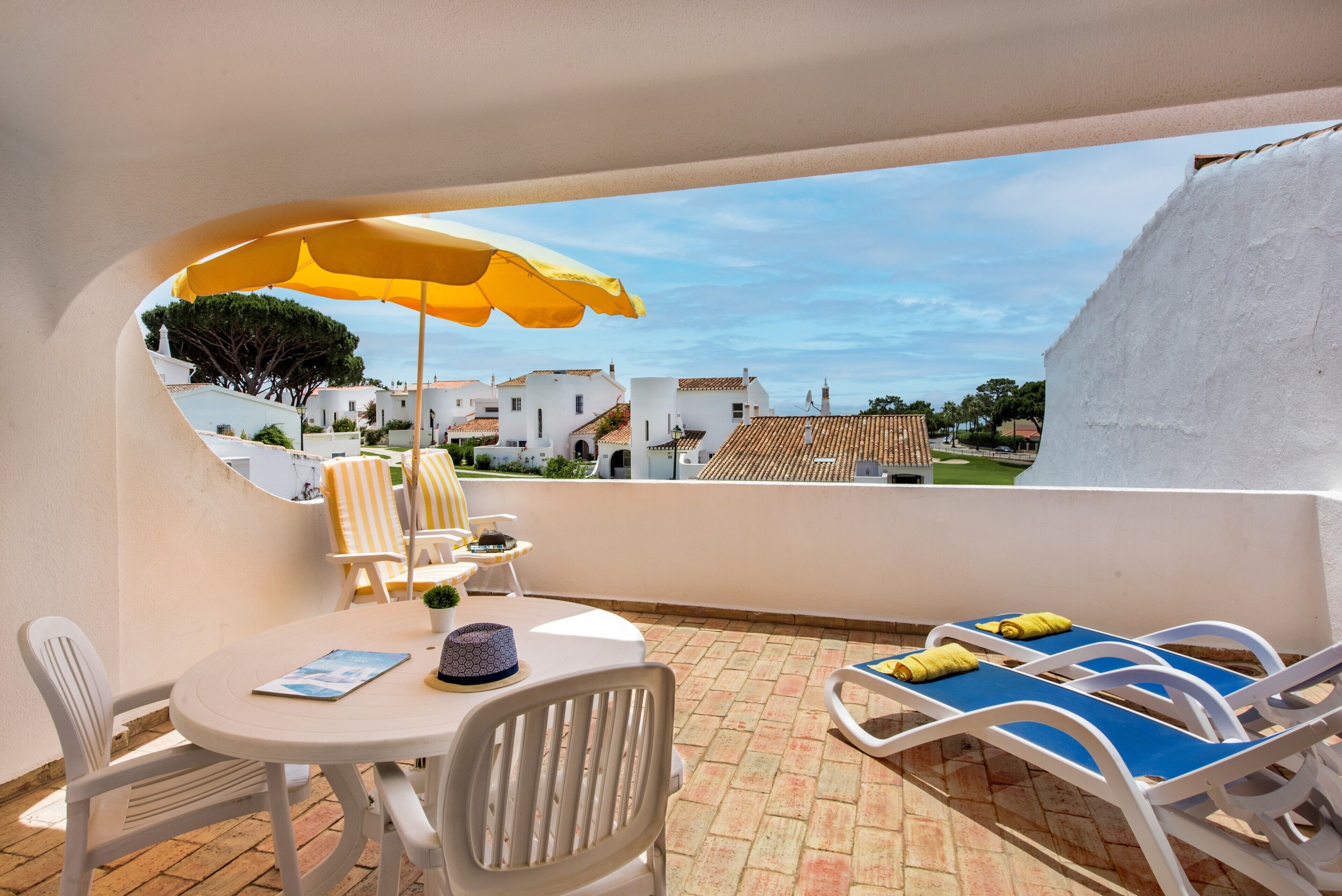 Un balcon avec des chaises blanches, une table et un parasol jaune.