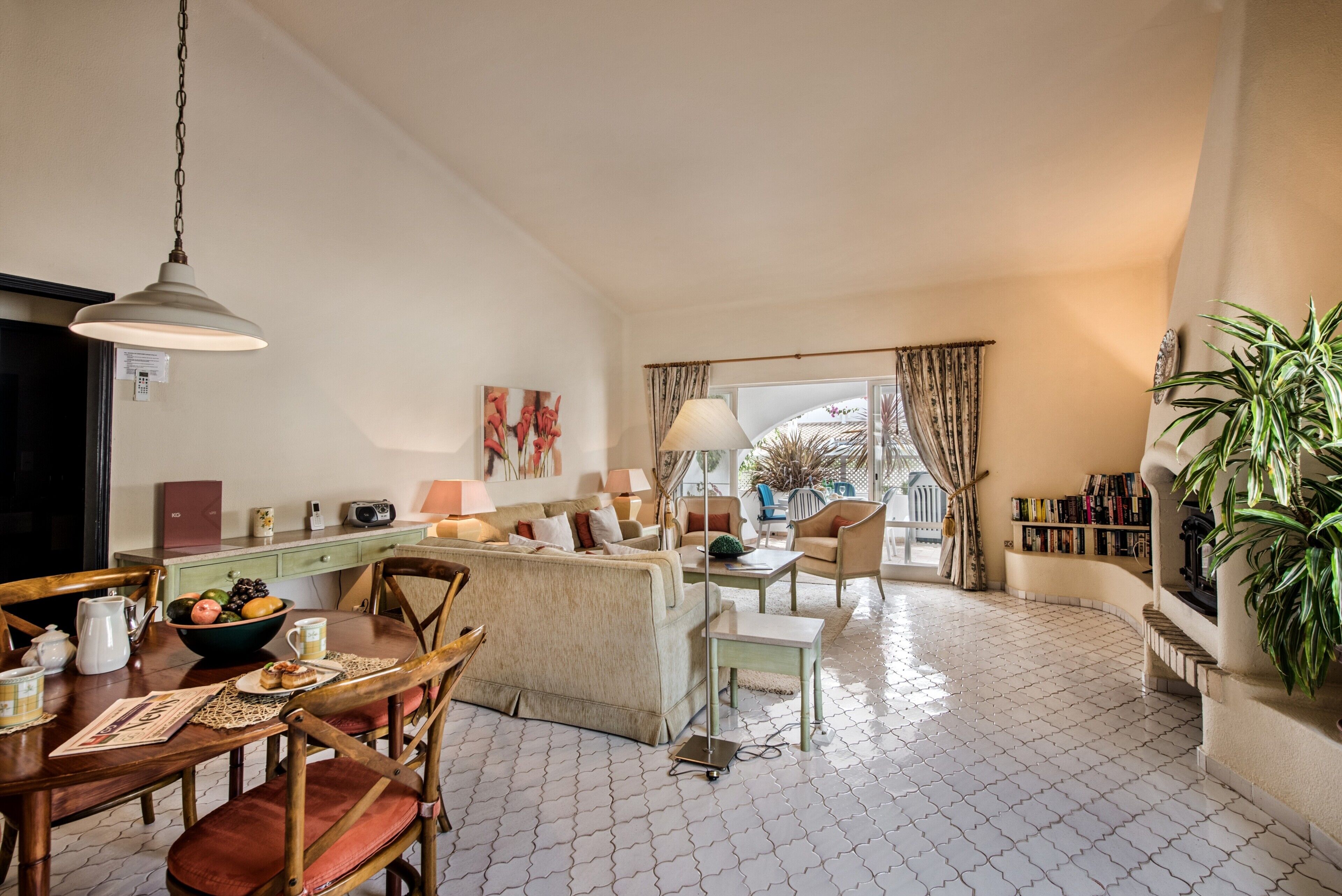 A spacious living area with a dining table, chairs, a sofa, and a coffee table. There is a window with curtains, a lamp, and a bookshelf.
