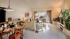 Amplia sala de estar con mesa de comedor, sillas, sofá y mesa de centro. Hay una ventana con cortinas, una lámpara y una estantería.