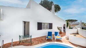 Un edificio blanco con piscina, dos sillones de descanso y un pequeño jardín.
