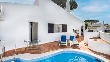 A white building with a swimming pool, two lounge chairs, and a small garden area.