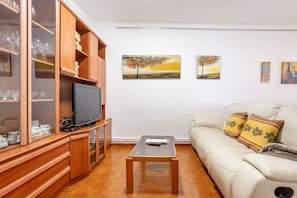 Una sala de estar con un televisor sobre un mueble de madera, una mesa de centro de vidrio, un sofá con almohadones decorativos y dos cuadros en la pared.