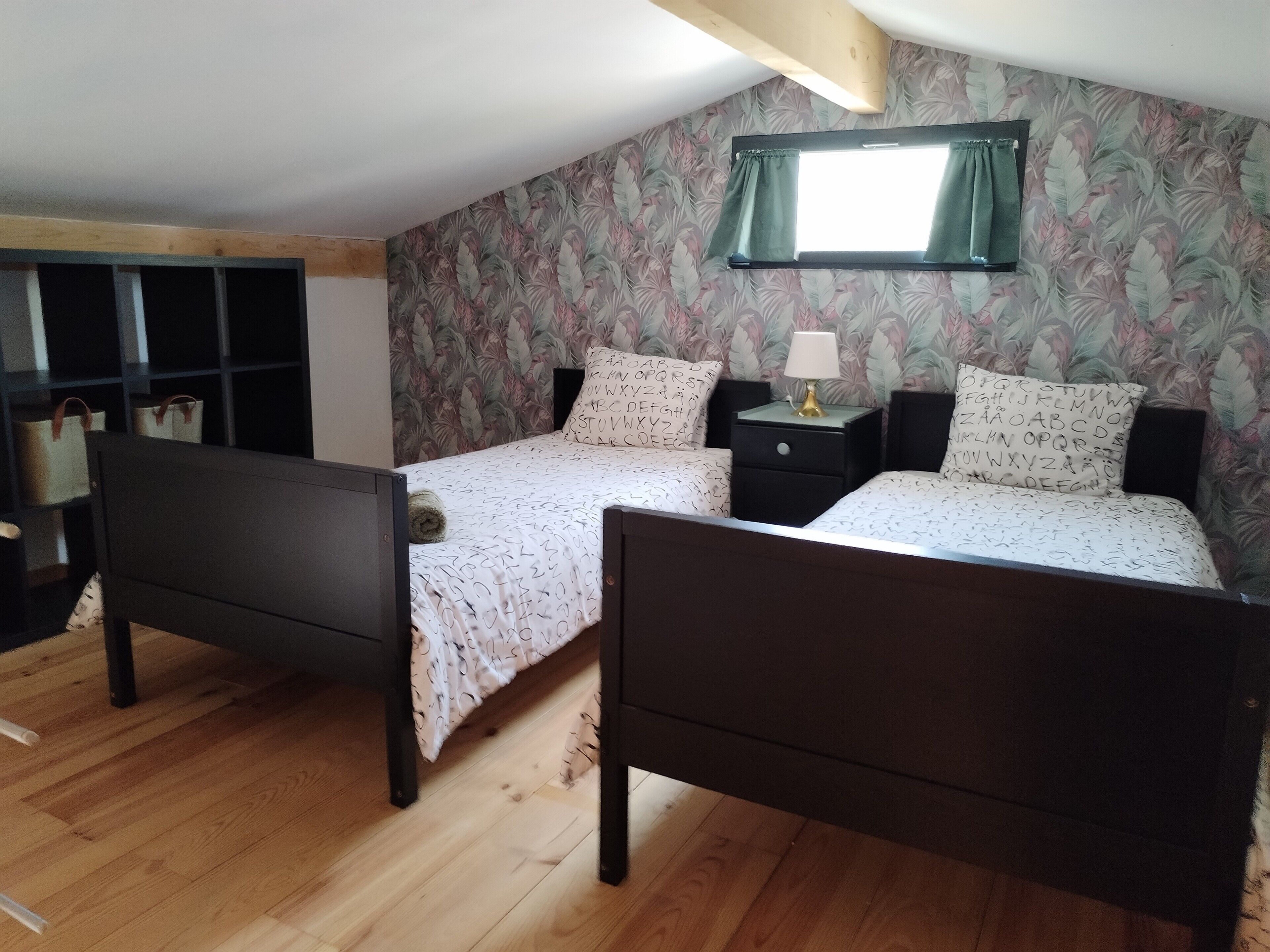 A bedroom with two beds, a wooden floor, a window with a curtain, and a wall with a leafy pattern.