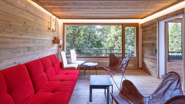 A wooden interior with a red sofa, leather chairs, a small table, and large windows offering a view of trees.