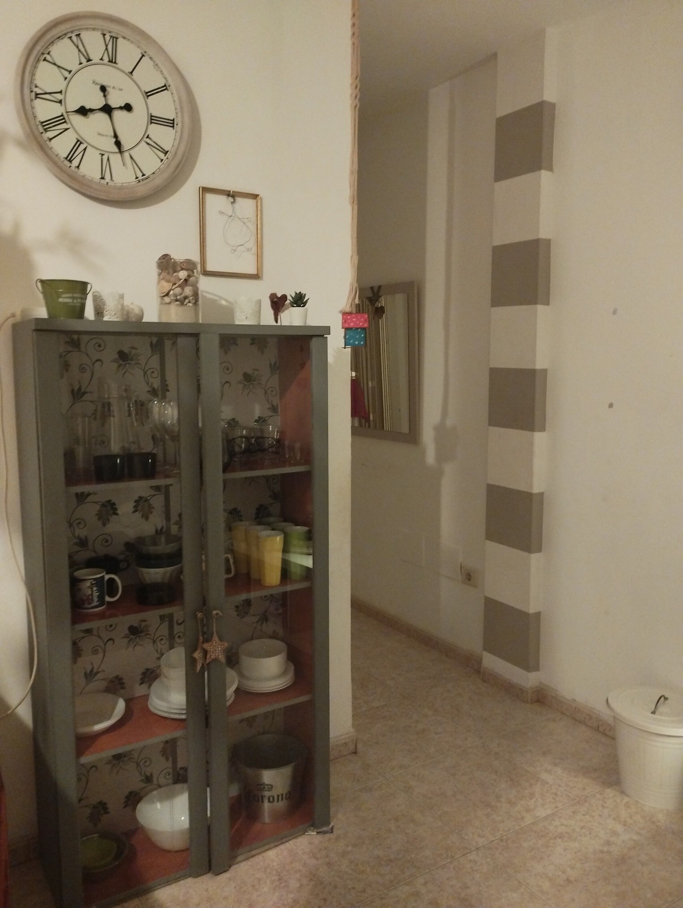 A room with a wall-mounted clock, a cabinet with dishes, and a hallway with a mirror.